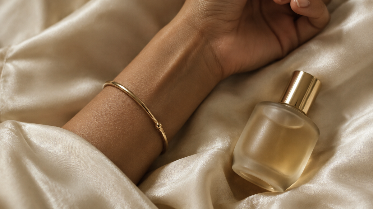 Wrist with gold bangle beside a small frosted perfume bottle on ivory silk